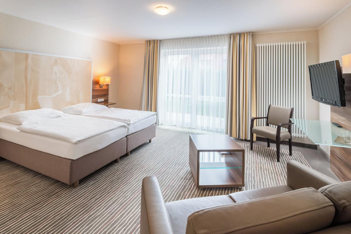 A hotel room with a large bed, a smaller bed, a sofa, a chair, a TV, and a window with curtains.