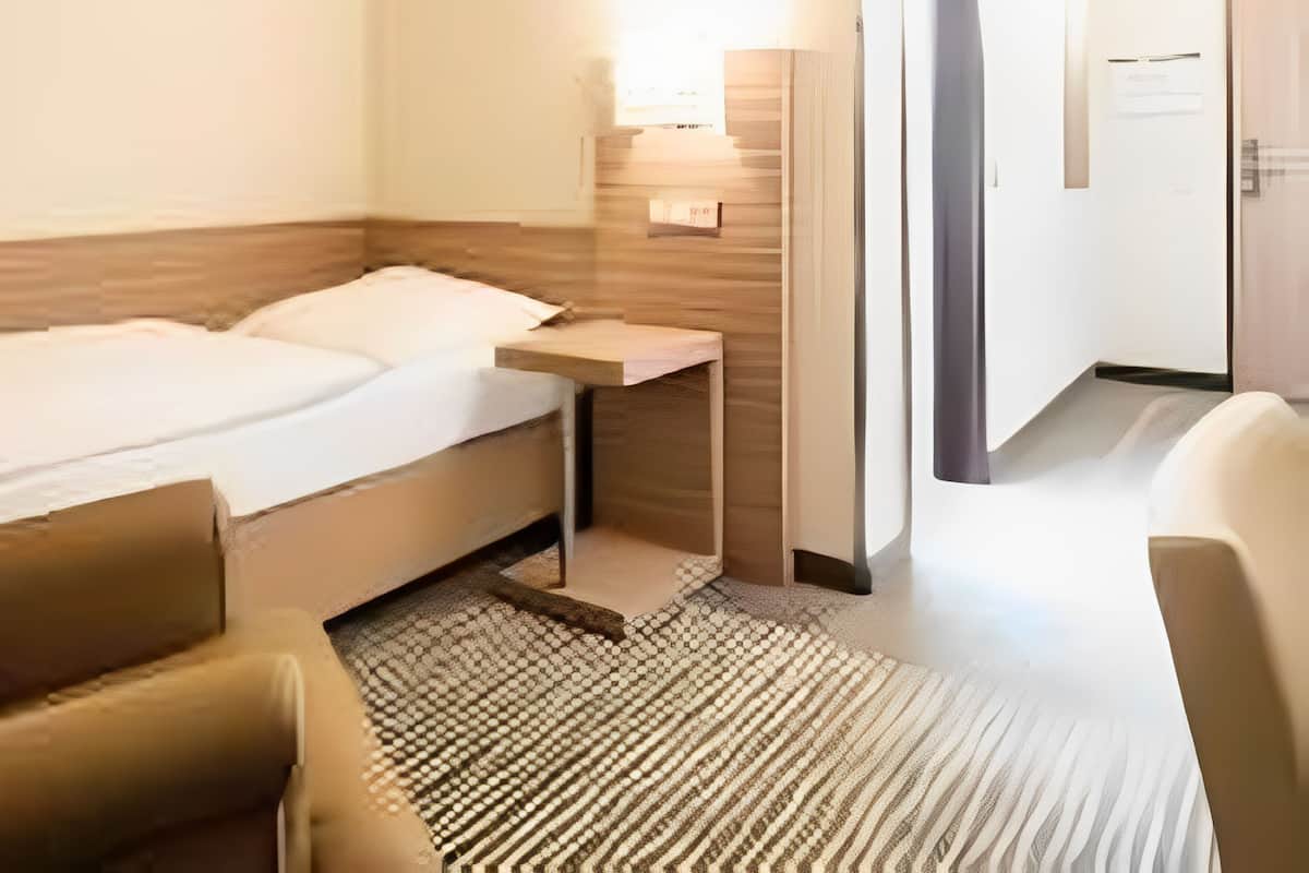 A hotel room with a bed, a bedside table, a lamp, a chair, and a door.