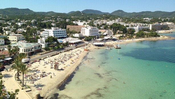 A coastal town with a sandy beach, clear turquoise waters, and a backdrop of green hills. - Hostal Ferrer (Sant Antoni de Portmany)