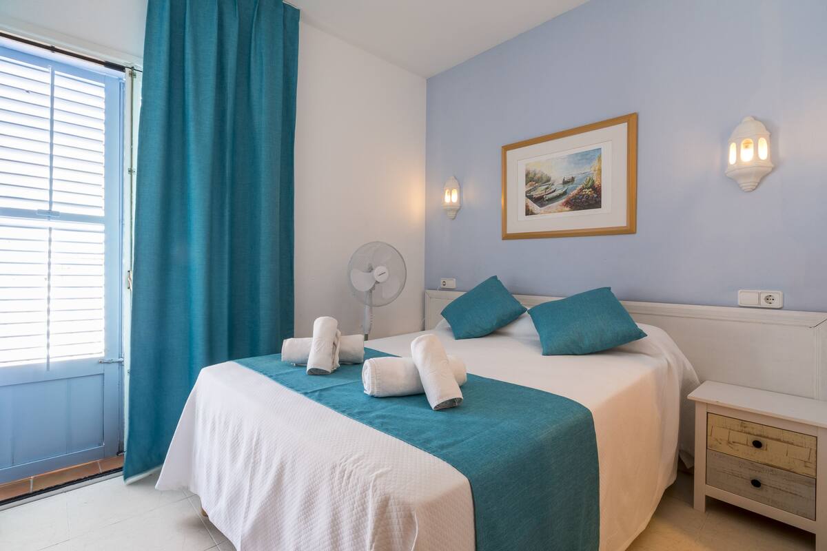 Ein ordentlich bezogenes Bett mit türkisfarbener und weißer Bettwäsche, ein Standventilator und eine Wandleuchte.