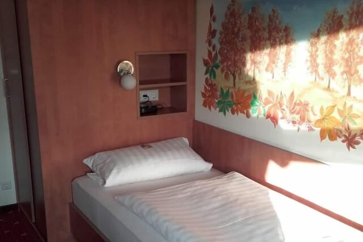 Una cama individual con ropa blanca en una habitación con pared roja y un mural de hojas otoñales.