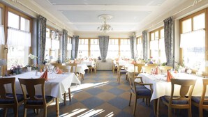 A spacious dining area with multiple tables and chairs, large windows, and a chandelier.