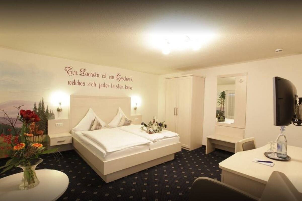 Habitación de hotel con cama, escritorio, silla, espejo y una pared con una frase decorativa.
