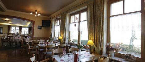 A dining area with wooden tables and chairs, large windows with curtains, and a television mounted on the wall.