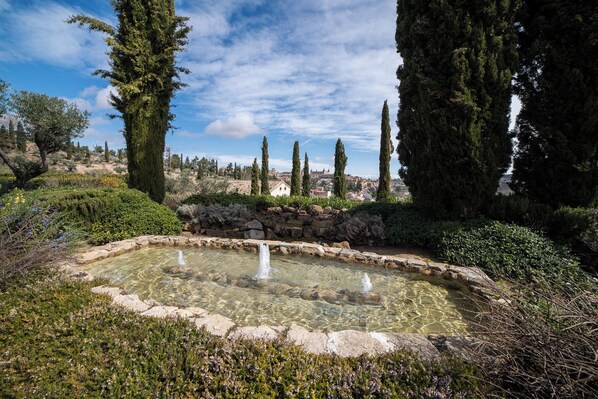 Una piscina al aire libre cristalina rodeada de muros de piedra y diversas plantas, con altos cipreses al fondo.