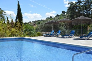 Una piscina con agua azul, rodeada de sillas de descanso y sombrillas, con zonas verdes y edificios al fondo.