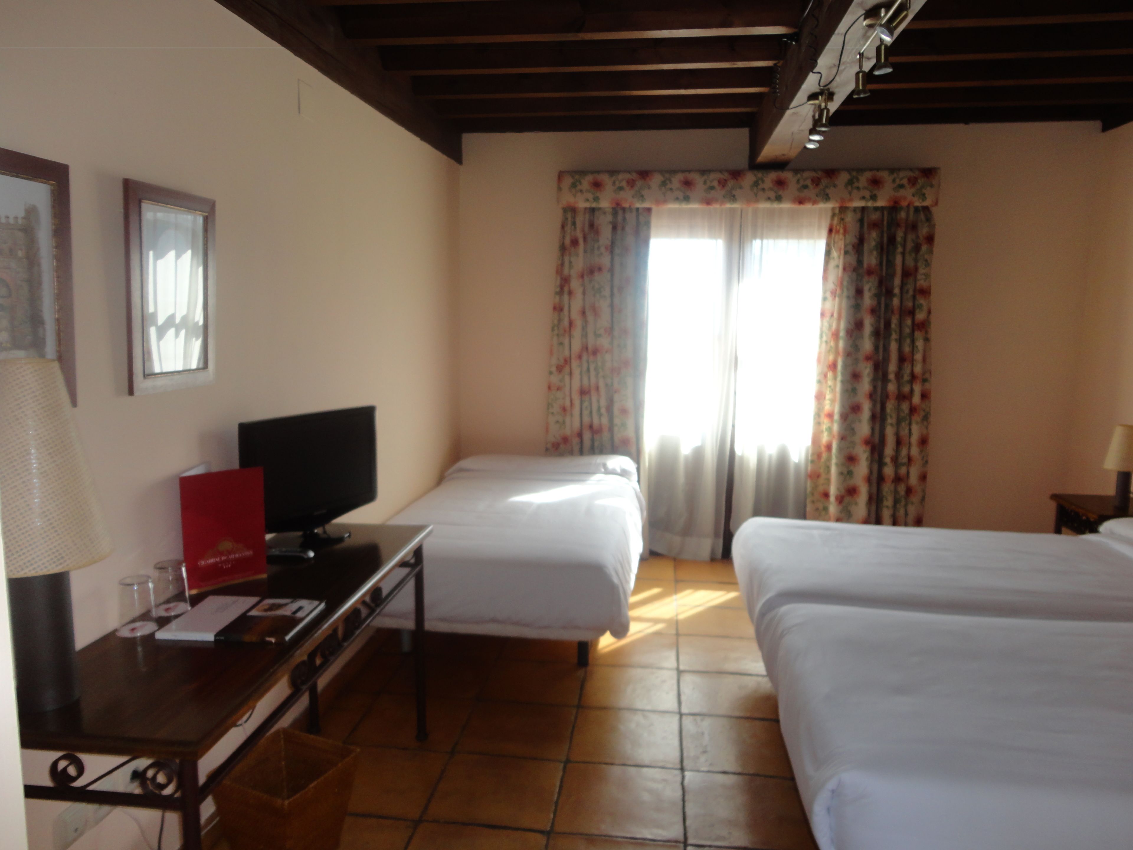 A hotel room with two beds, a TV on a desk, and a window with floral curtains.
