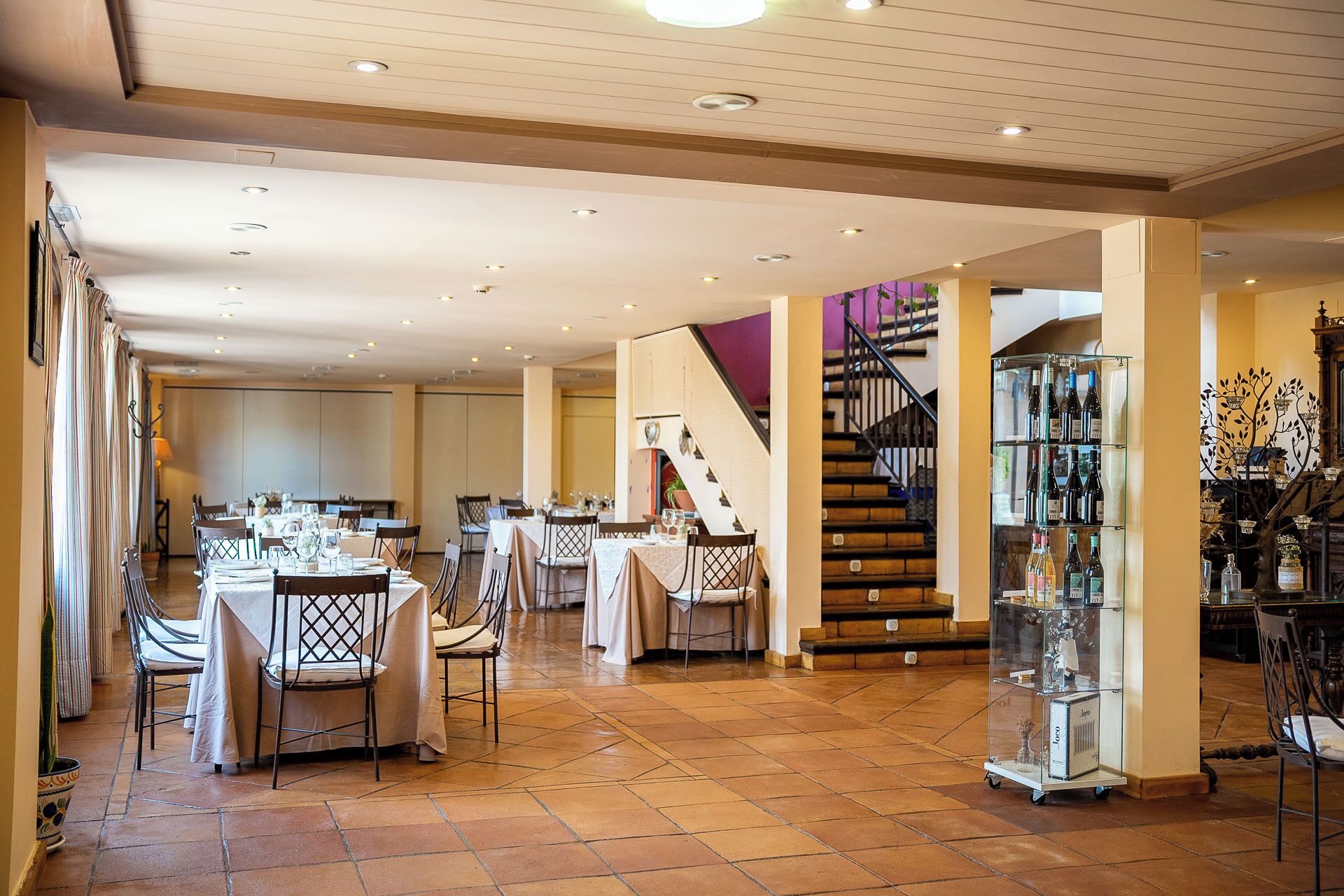 Un comedor amplio con varias mesas y sillas, una escalera y una vitrina de vinos.