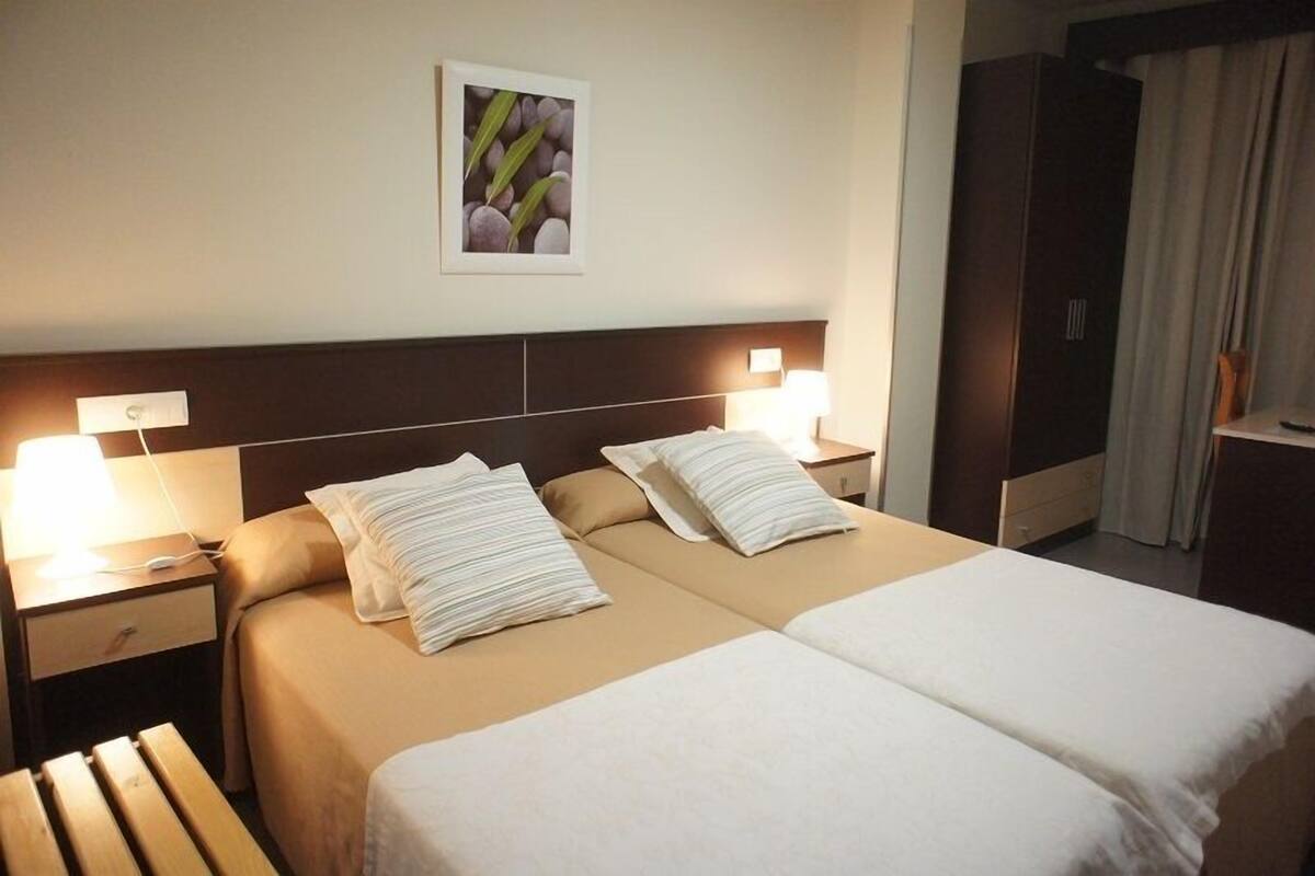 A hotel room with a double bed, two bedside tables with lamps, a nightstand, a wooden bench, and a wardrobe.