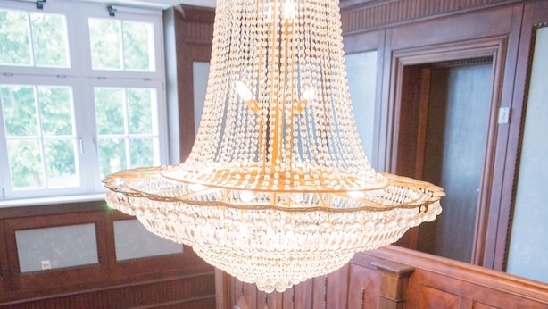 A grand hall with a large chandelier, wooden paneling, and a desk with chairs. - Villa Ingeborg (Fürstenberg-Havel)