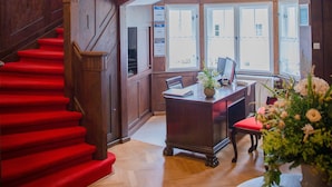 A grand staircase with red carpet leading to a second floor, wooden paneling, and a desk with a vase of flowers. - Villa Ingeborg (Fürstenberg-Havel)