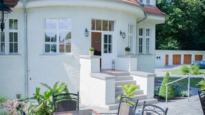 A patio with a wooden table and wicker chairs, a white building with a red roof, and a small garden area with potted plants. - Villa Ingeborg (Fürstenberg-Havel)