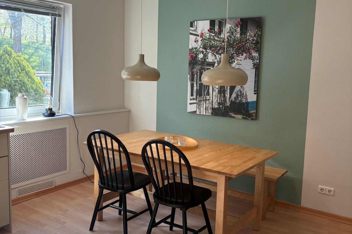 A dining area with a wooden table, two black chairs, a hanging pendant light, and a wall-mounted picture.