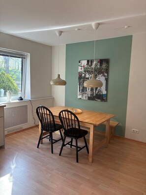 Uma sala de jantar com uma mesa de madeira, duas cadeiras pretas, uma luminária pendente e um quadro na parede.