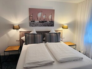 A hotel room with a bed, two nightstands, a painting of sailboats, and two lamps. - Hotel Haus am See (Sinzheim)
