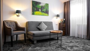 Een moderne hotelkamer met een grijze bank, een bijpassende fauteuil en een klein houten bijzettafeltje. Twee wandlampen, een schilderij van een boslandschap en een raam met gordijnen.
