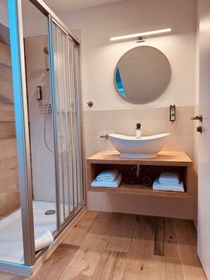 A modern bathroom with a glass shower enclosure, a round mirror, and a wooden vanity with a white sink. - Hotel Haus am See (Sinzheim)