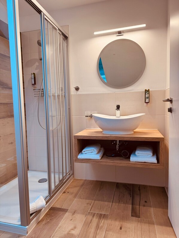 Une salle de bain moderne avec une douche à parois de verre, un miroir rond et un meuble-lavabo en bois avec un vasque blanche.