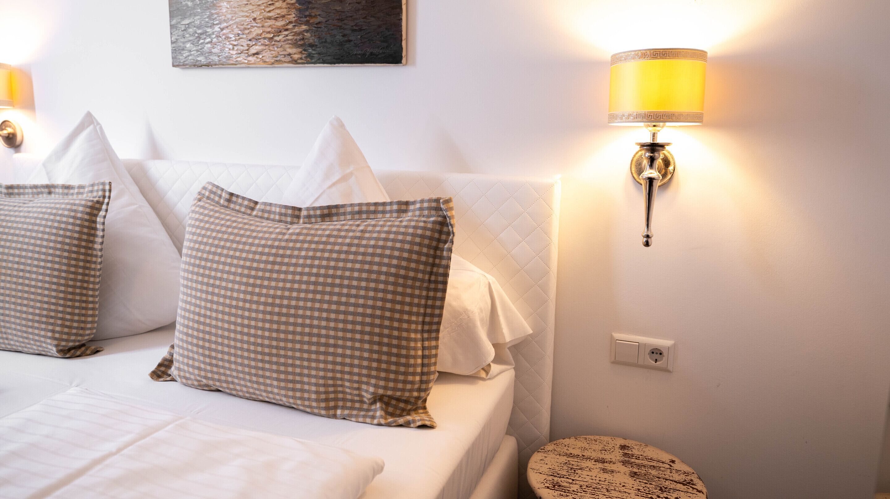Una cama bien hecha con almohadones a cuadros, una lámpara de noche y una pequeña mesa redonda.