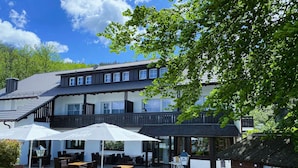 Een gebouw met twee verdiepingen, een balkon, een terras met tafels en parasols, en een verzorgde tuin.