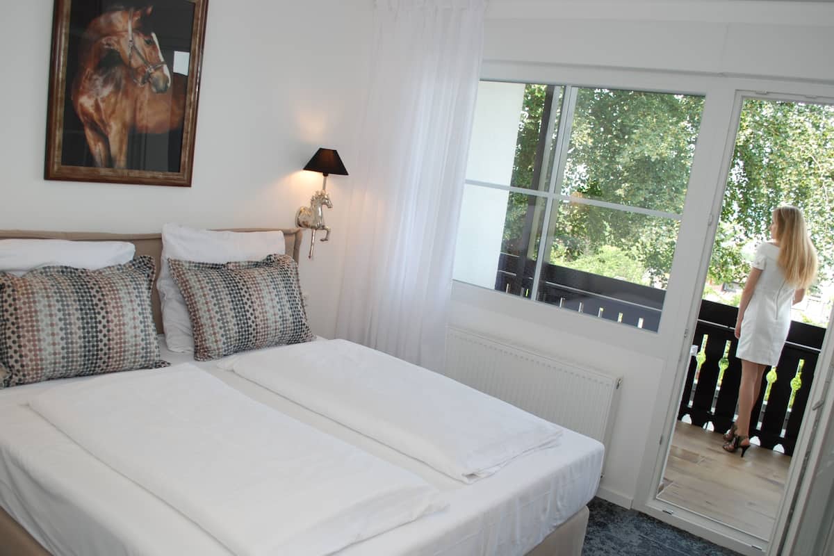 A bedroom with a large bed, a nightstand, a lamp, a window with a view of greenery, and a balcony with a person standing.
