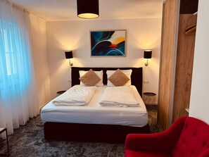 A hotel room with a bed, two wall-mounted lamps, a painting, a red chair, and a wooden wardrobe. - Hotel Haus am See (Sinzheim)