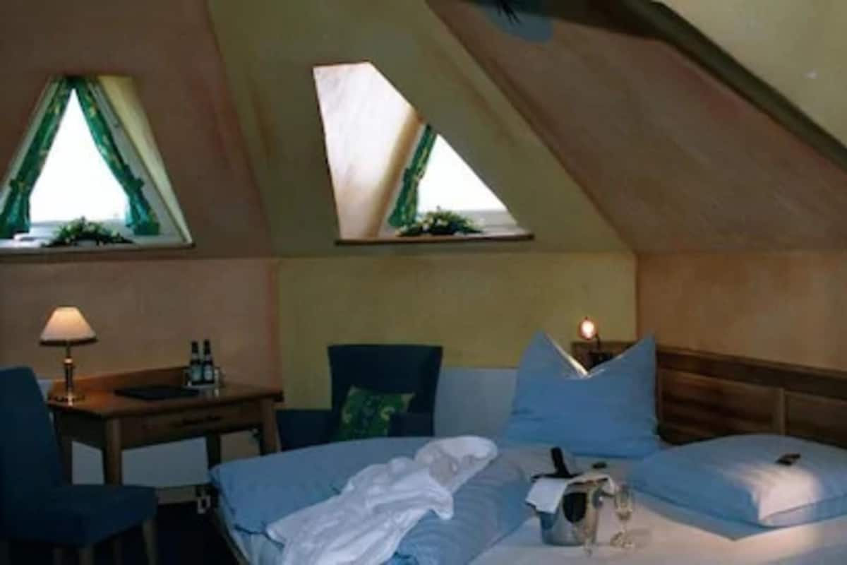 Una habitación pequeña y acogedora con techo inclinado, cama con ropa de cama azul, una mesita de noche con lámpara y una silla.