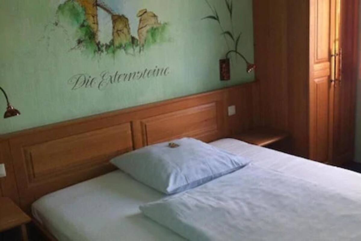 Un dormitorio con cabecera de madera, una cama individual con ropa de cama blanca y un mural en la pared con el texto "Die Externsteine".