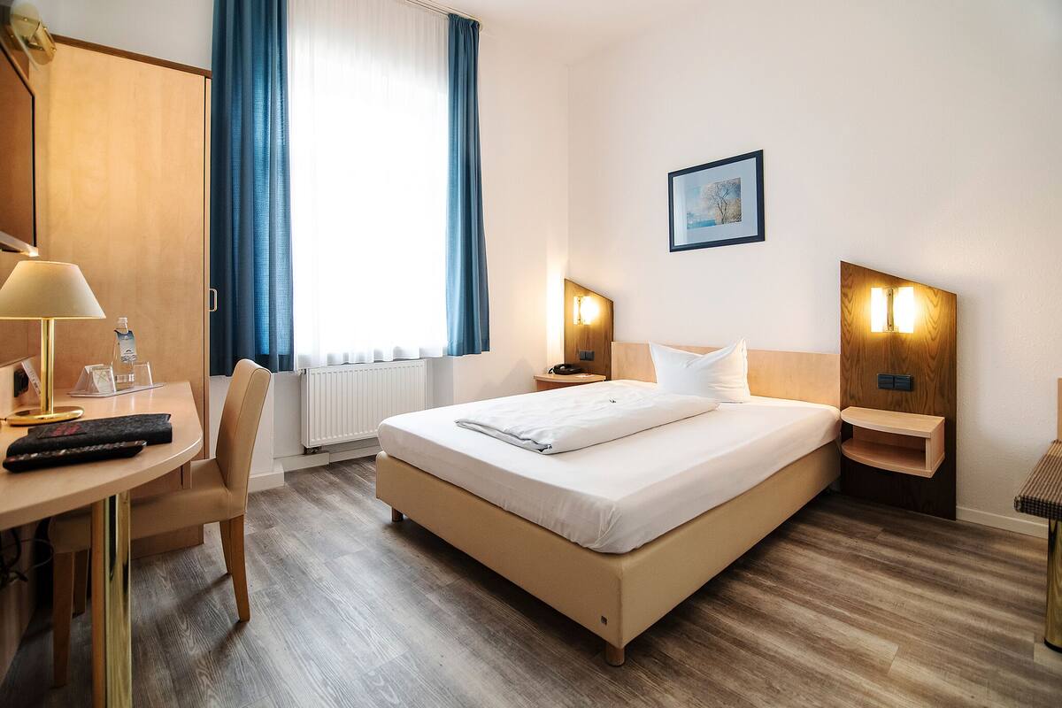 Een hotelkamer met een bed, nachtkastjes, een bureau met lamp, een stoel en een raam met gordijnen.
