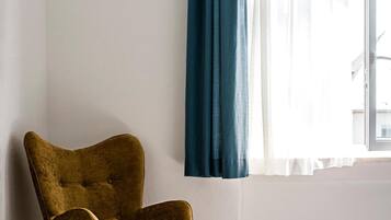 Una habitación con un sillón individual color mostaza, una ventana con cortinas blancas traslúcidas y un cortinero azul oscuro, y piso de madera.