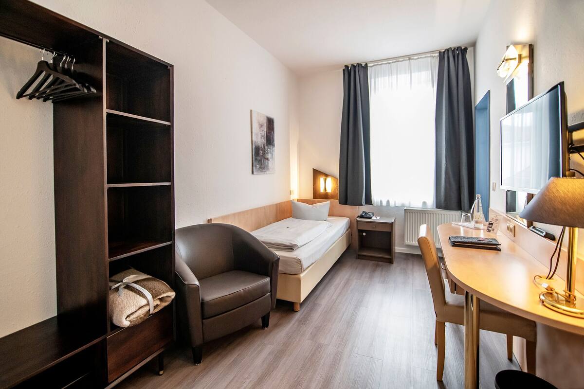 Een kleine hotelkamer met een bed, bureau, stoel, kledingkast en raam met gordijnen.