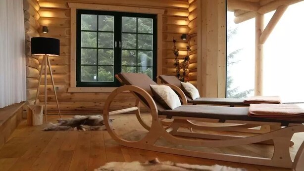 A log cabin interior with a wooden ceiling, a large window, a lounge chair, a floor lamp, and a fur rug.