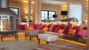 A log cabin interior with a stone fireplace, a sofa with colorful cushions, and plaid-patterned ottomans.