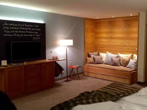 Una habitación acogedora con pared de madera, un televisor sobre un mueble de madera, un sofá con cojines y una mesita con una lámpara.