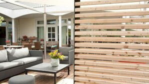 A wooden deck with a sofa and a table, a potted plant, and a view of a house with a covered patio. - Golfhotel Gut Winterbrock (Rheine)