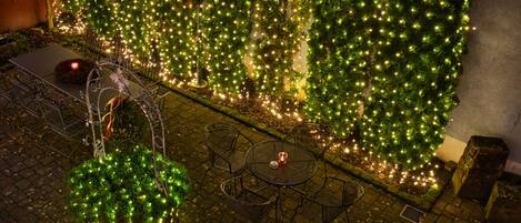 Un área de estar al aire libre con una mesa y sillas, rodeada por un muro decorado con follaje verde y luces colgantes.