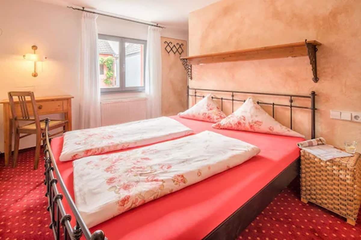 A double bed with floral bedding, a wooden desk, a chair, a small wicker side table, and a window with curtains.