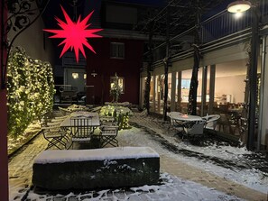 An outdoor seating area with snow-covered tables and chairs, a stone bench, and a Christmas tree with lights.