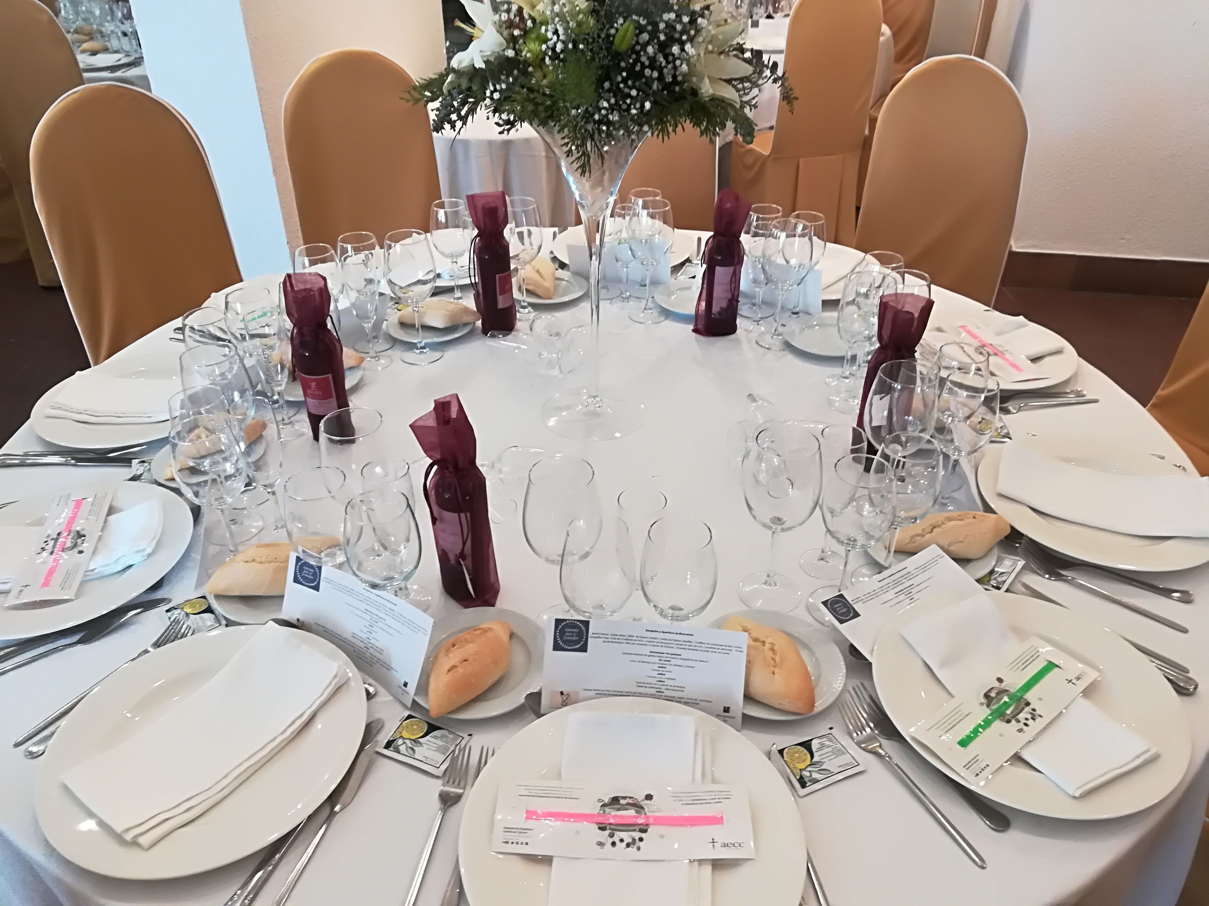 Una mesa redonda para comer servida con platos blancos, copas de vino transparentes y servilletas dobladas.