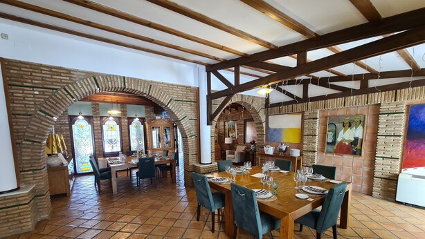 A spacious dining area with a high ceiling and exposed wooden beams, featuring a rustic brick wall and tiled flooring.