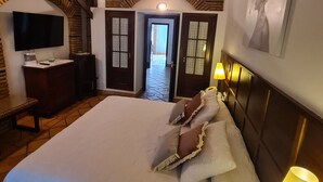 A spacious bedroom with a large bed, a TV mounted on a brick wall, a desk with a lamp, and a door leading to a hallway.