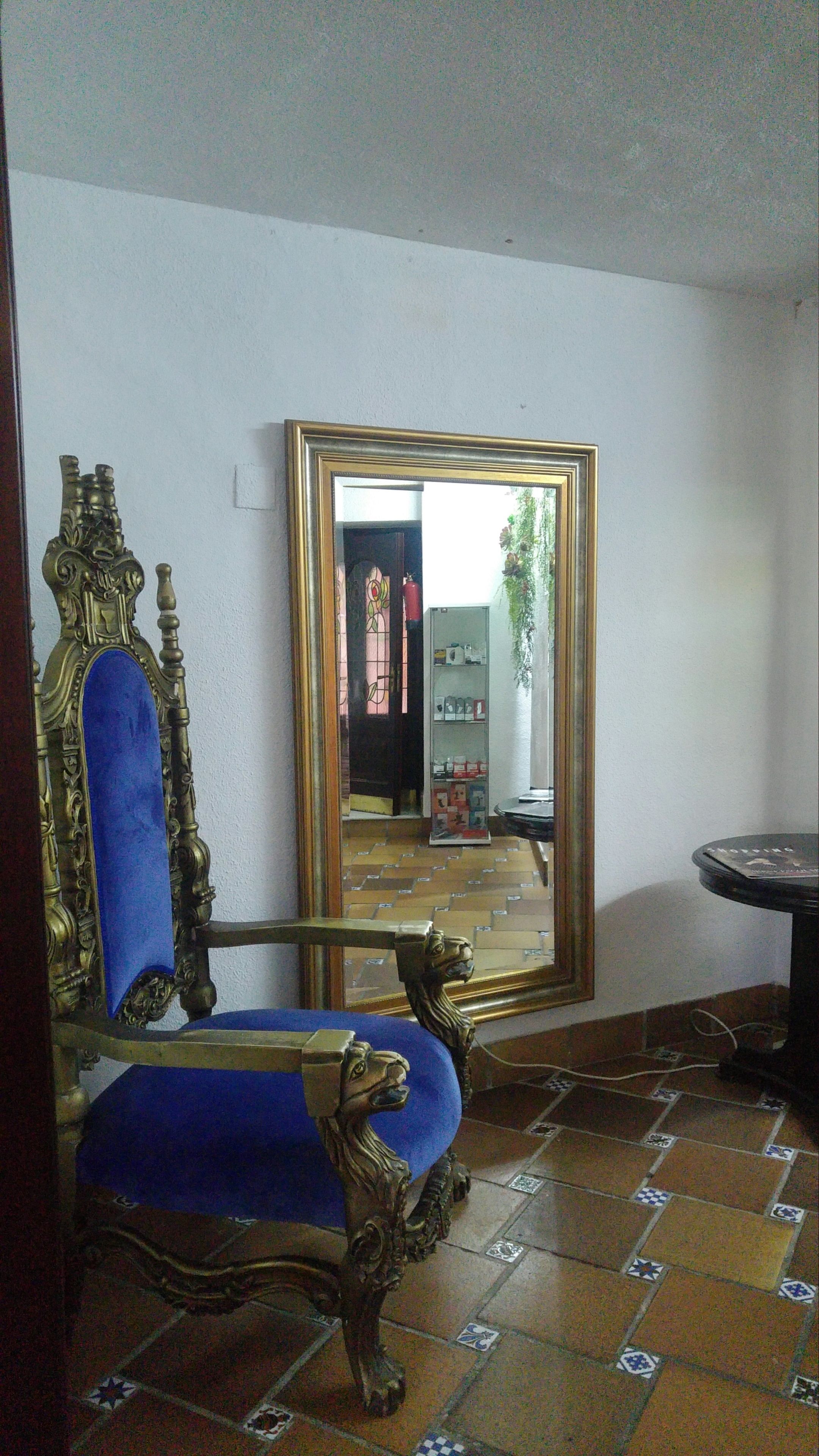 Una habitación con un espejo grande y decorativo, un sillón tapizado en azul y un suelo de baldosas.