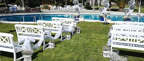 Zona junto a la piscina con sillas y mesas blancas, cielo azul despejado y follaje alrededor.