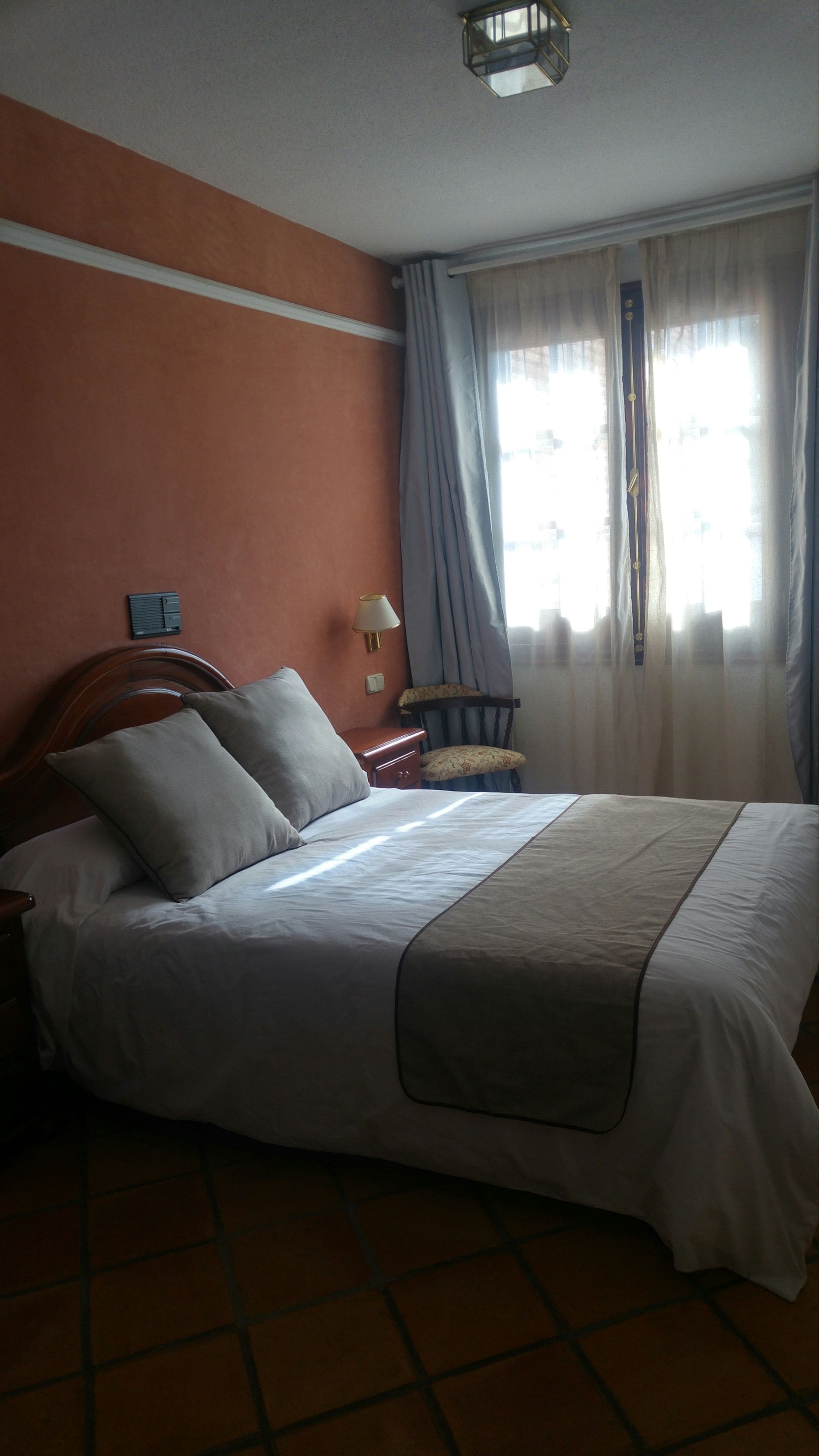 A bedroom with a bed, a nightstand, a chair, a lamp, and a window with curtains.