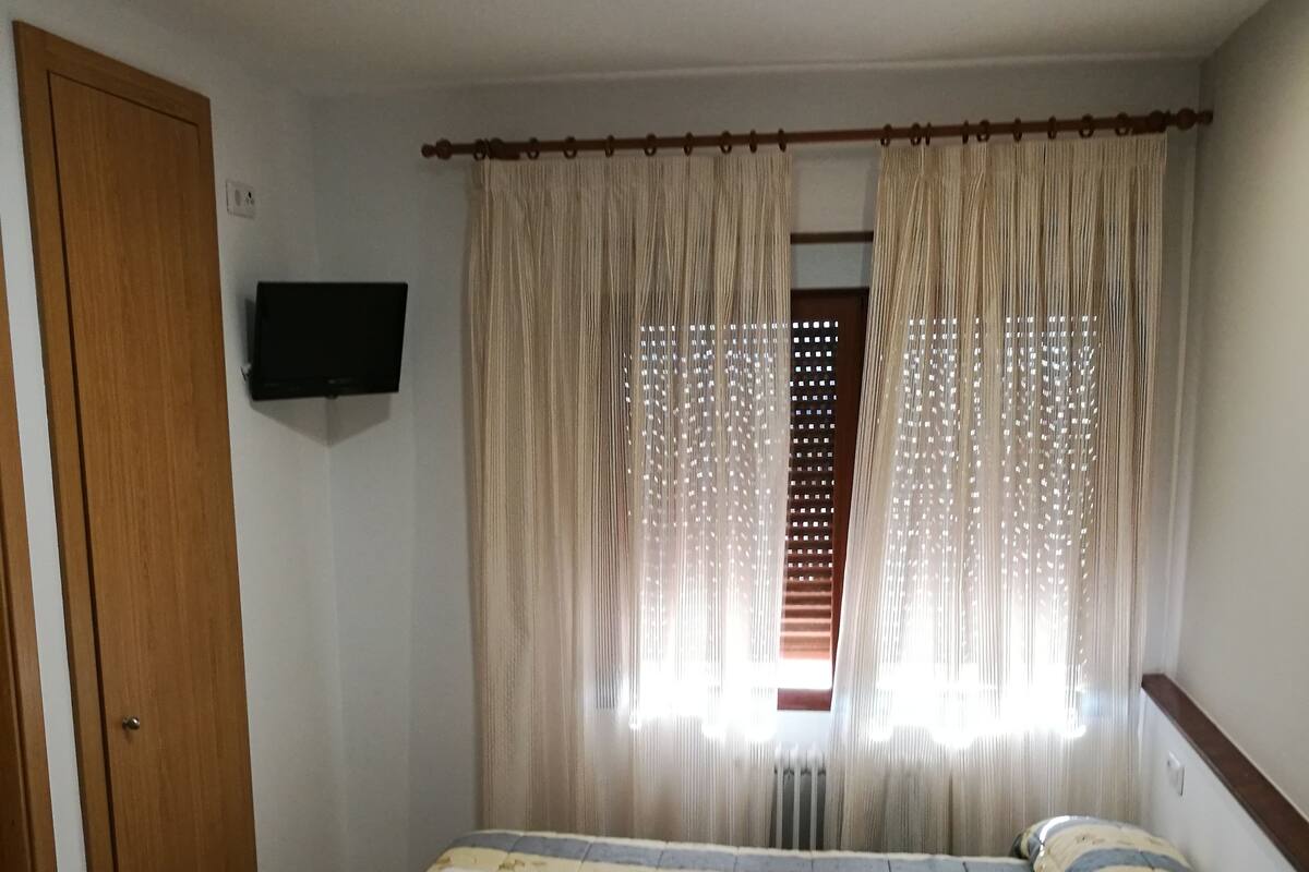 A room with two beds, a wooden door, a television mounted on the wall, and a window with curtains.