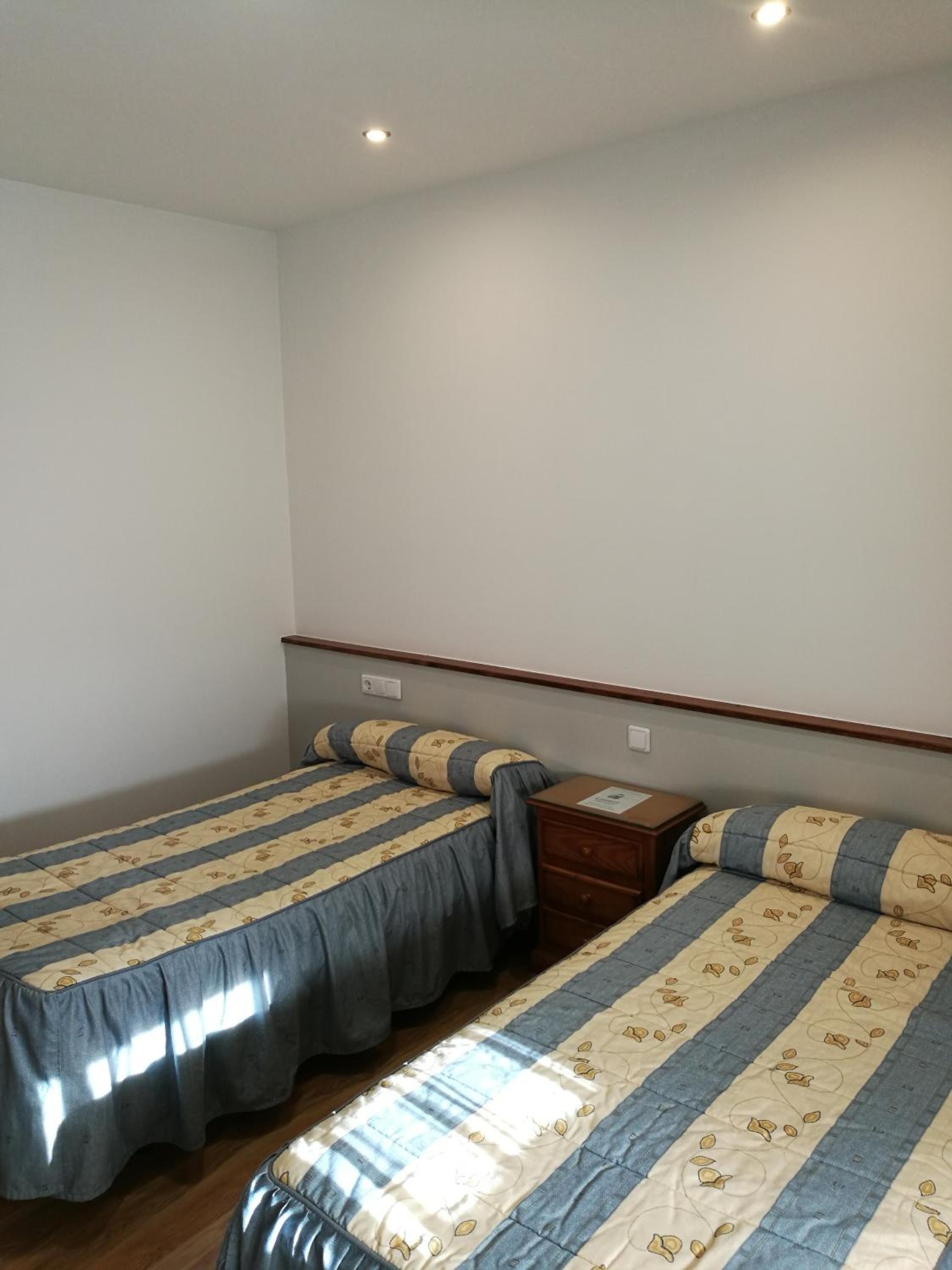 Una habitación pequeña y limpia, con dos camas, una mesita de noche y un aparador de madera.