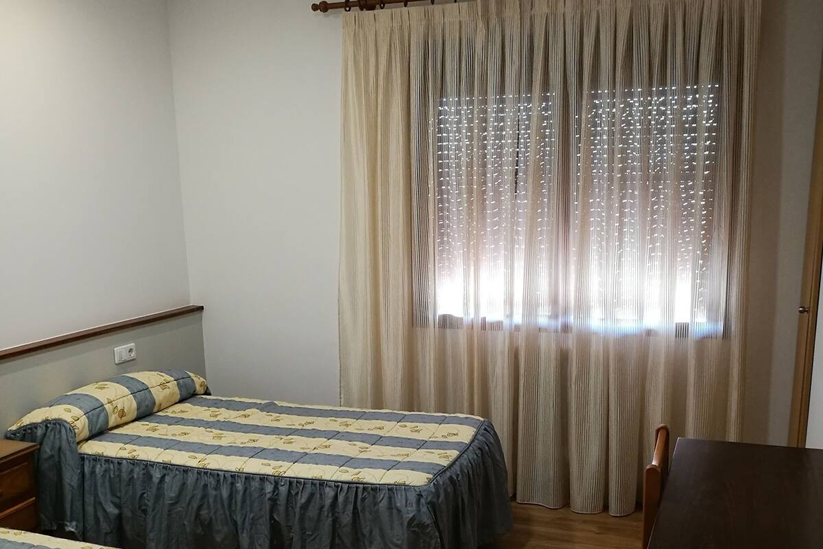 A room with two beds, a wooden table, and a window with curtains.