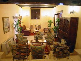 A cozy living room with patterned armchairs, a sofa, and a coffee table. There are potted plants and a wooden cabinet.