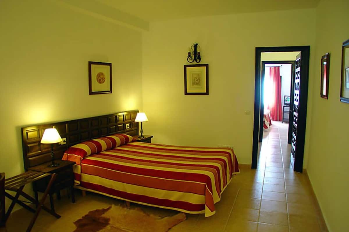 A hotel room with a large bed, two bedside lamps, a wooden headboard, a framed picture on the wall, and a door leading to another room.