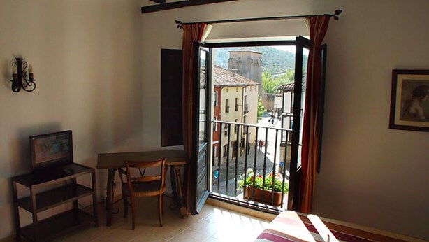 A room with a bed, a desk, a chair, and a balcony with a view of buildings and a mountain. - Nuevo Arlanza (Covarrubias)